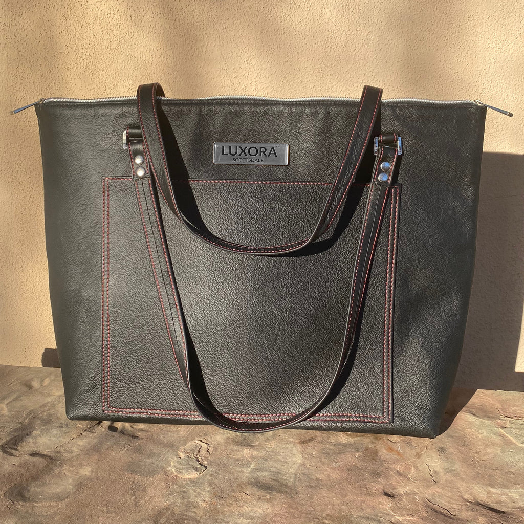 Black leather tote bag with visible 'Luxora' brand with red stitching on brown stone surface